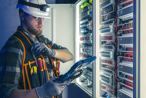 Electrician focused on work in switchboard with fuses, using tablet. Young adult electrical engineer in special clothes with flashlight on helmet in dark room with emergency lights in background.